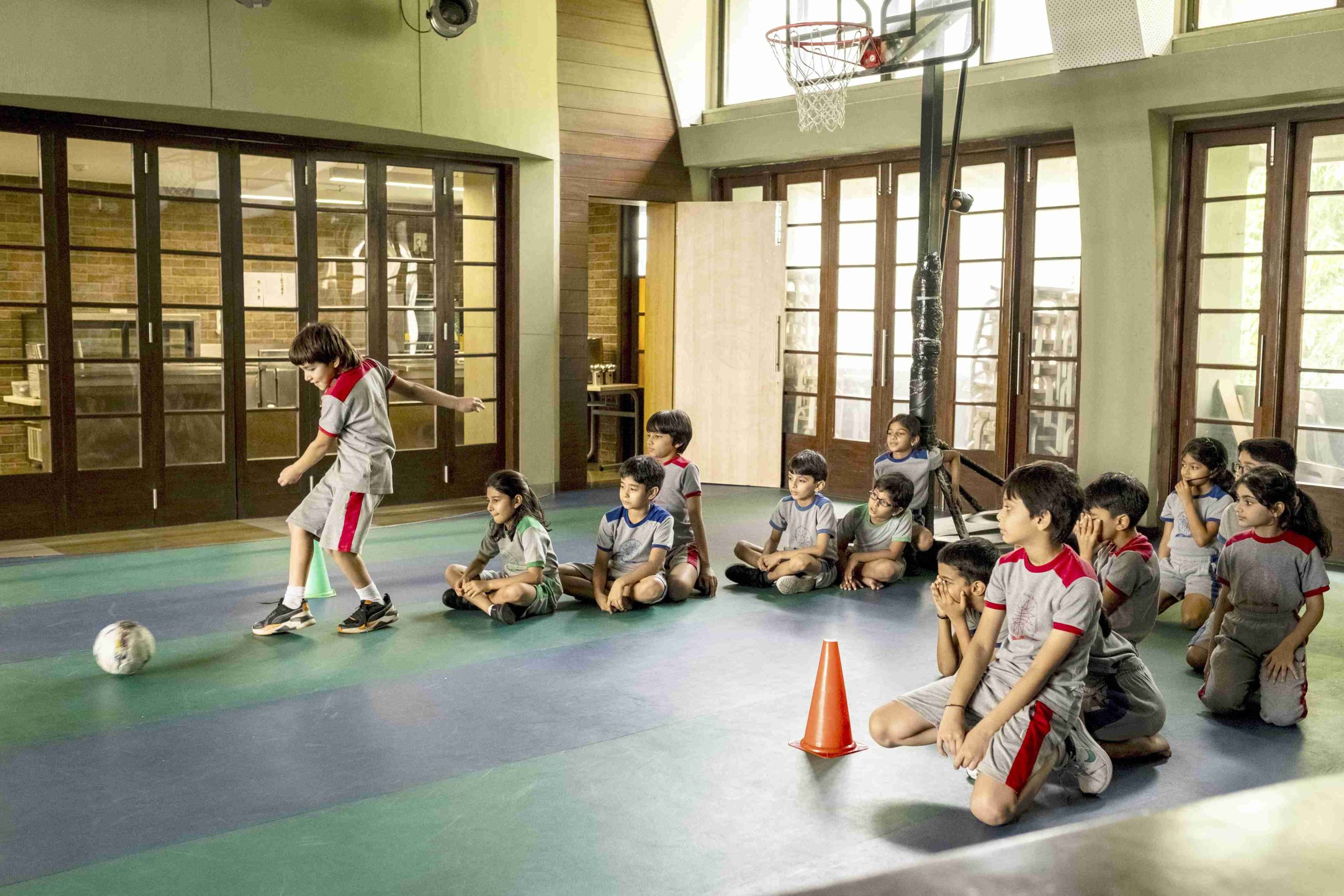 a kid is kicking ball while other sitting behind in an indoor facility